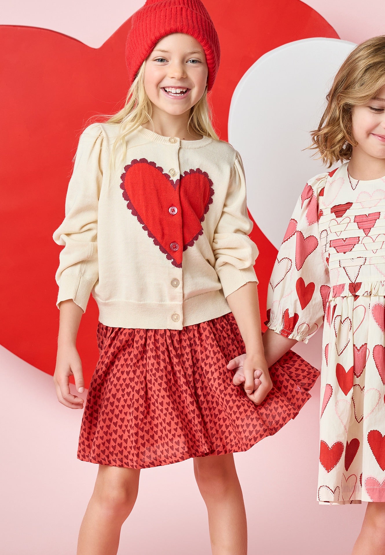 Two young girls wearing Pink Chicken Girls' Gianna Skirts and heart print Valentine’s Day outfits smile in front of a pink and red heart backdrop.