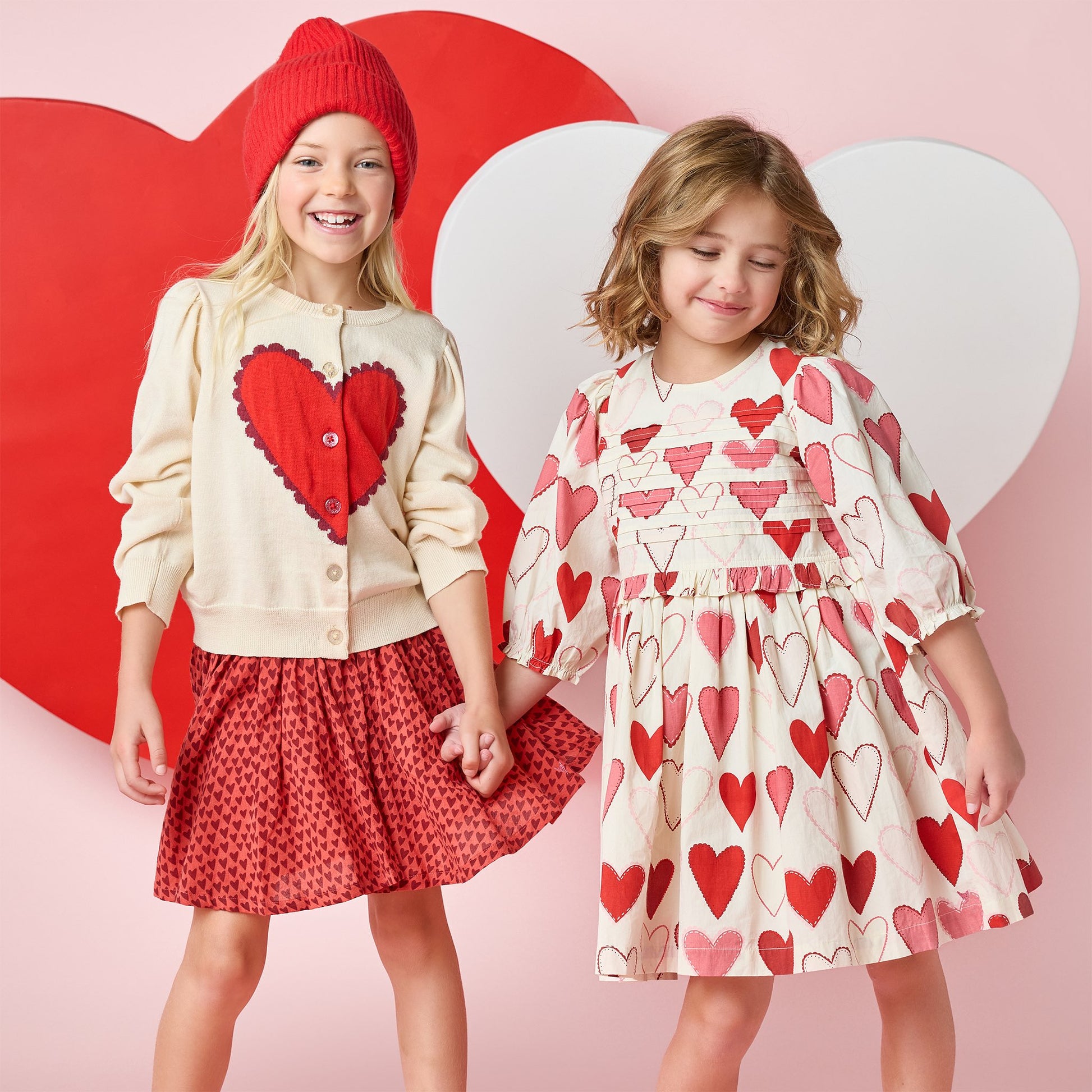 Two young girls smile and hold hands in Pink Chicken Girls' Renee Dresses with gradient hearts, posing before red and white heart-shaped backdrops—festive outfits perfect for Valentine’s Day.