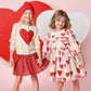 Two girls hold hands and smile in front of red and white heart-shaped backgrounds, both wearing matching Pink Chicken Girls' Constance Sweaters.