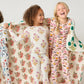 Three children wearing Pink Chicken Kids' Bamboo PJ Sets in cherry print smile together, holding a cream quilt with pink and green florals, set against a plain white background.