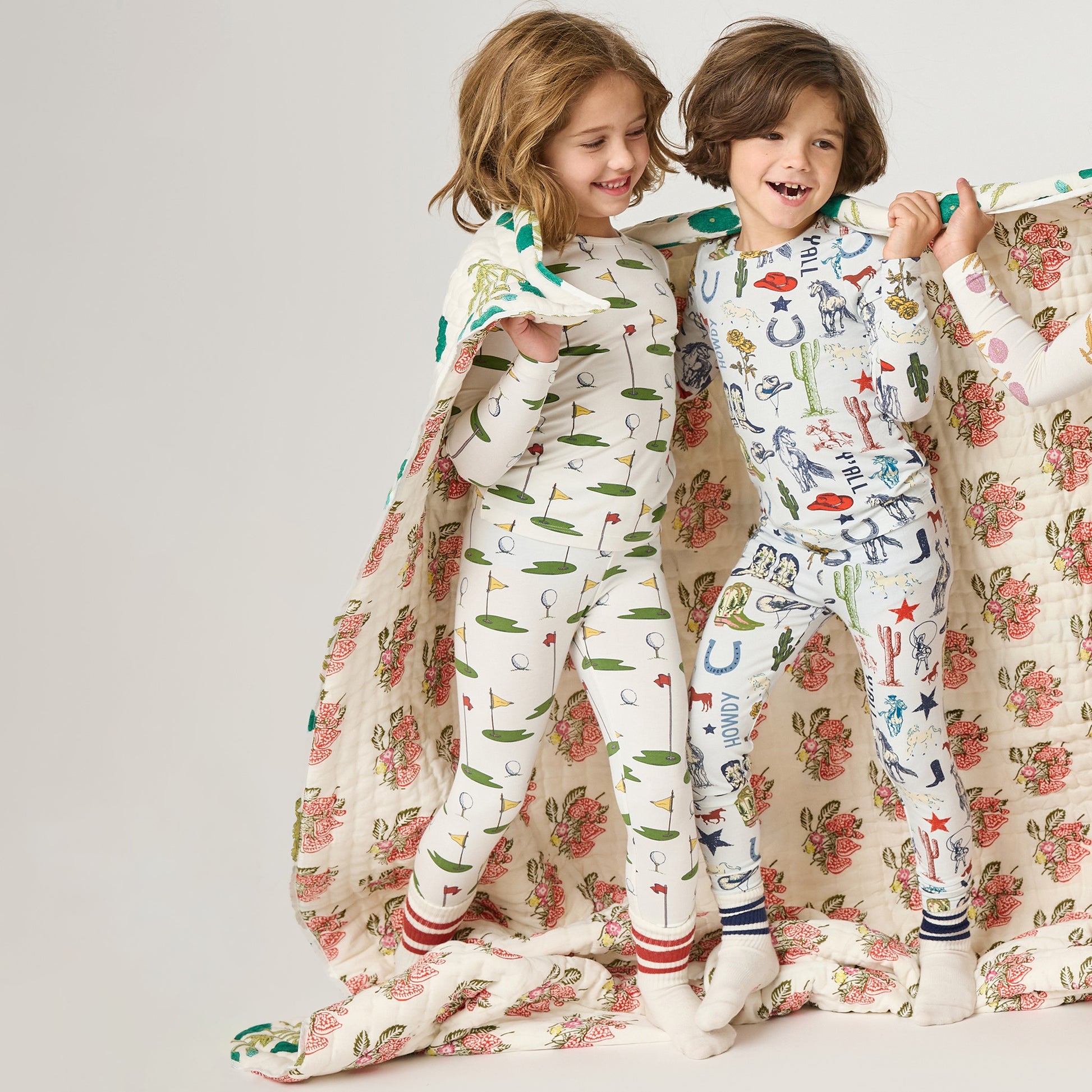 Two children wearing Pink Chicken Kids' Bamboo PJ Set with cherry print smile together, wrapped in a floral blanket against a plain background.