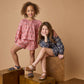Two young girls smile for the camera, one standing and one sitting on wooden blocks. Both wear Pink Chicken Girls' Theodore Short with matching pink tops and sandals, all set against a beige background.
