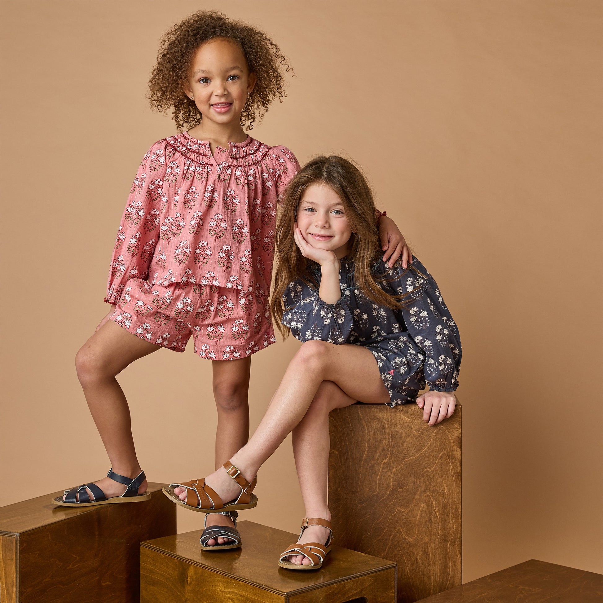 Two young girls smile for the camera, one standing and one sitting on wooden blocks. Both wear Pink Chicken Girls' Theodore Short with matching pink tops and sandals, all set against a beige background.
