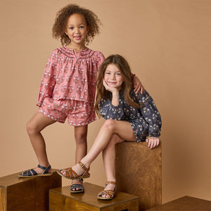 Two young girls smile for the camera, one standing and one sitting on wooden blocks. Both wear Pink Chicken Girls' Theodore Short with matching pink tops and sandals, all set against a beige background.