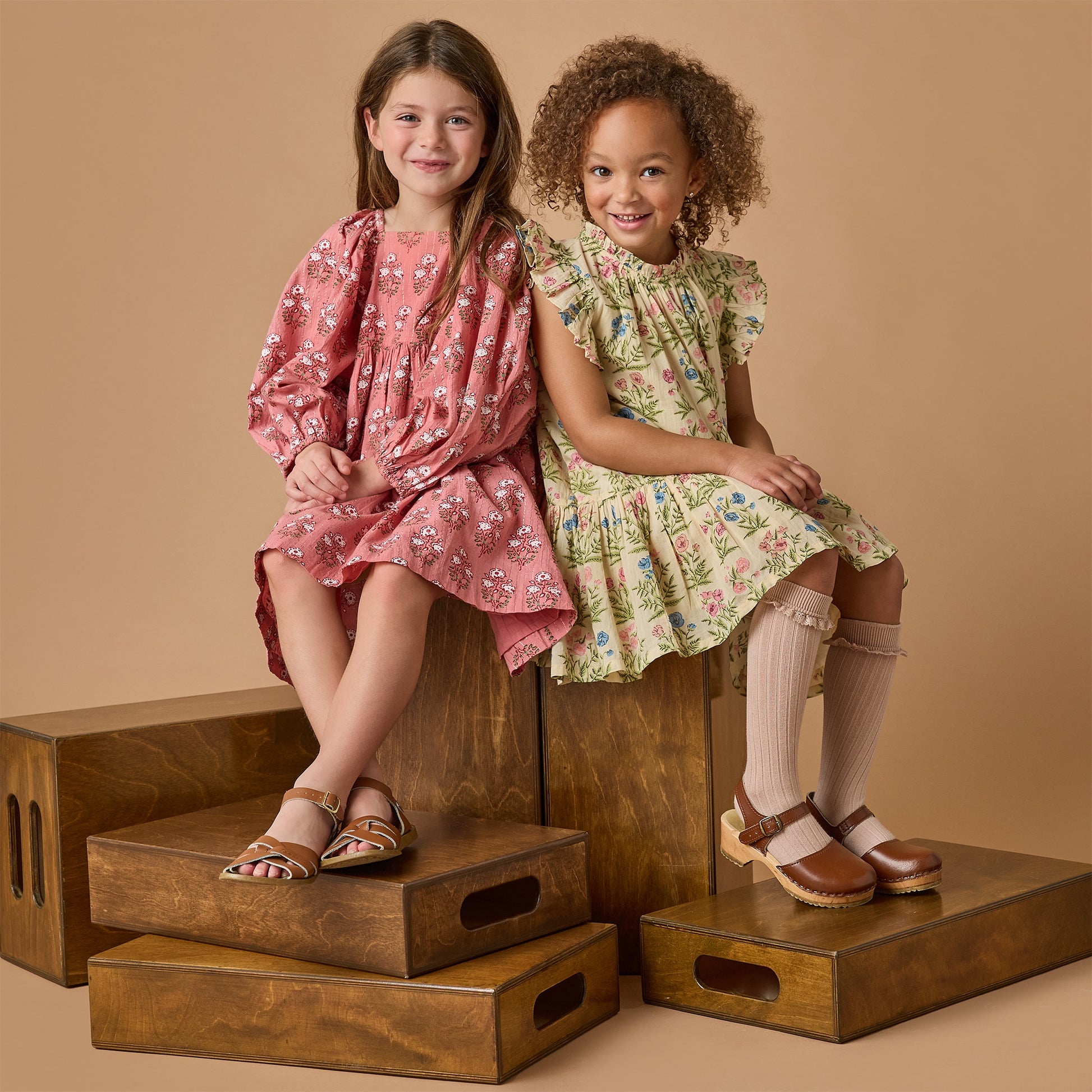 Two young girls sit on wooden boxes in a studio, both smiling in their sandals. One wears the Pink Chicken Girls' Mary Barrett Dress against a beige background.