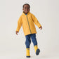 A young child stands against a plain background, smiling and lifting one foot slightly off the ground while wearing blue pants, a yellow raincoat, and Hatley Yellow & Navy Matte Rain Boots.