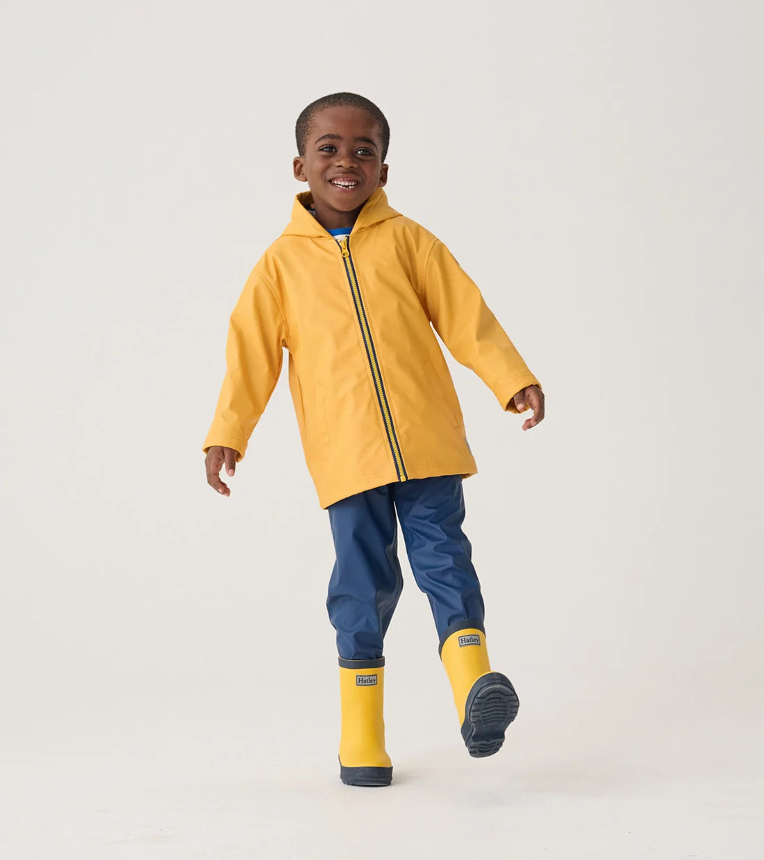 A young child stands against a plain background, smiling and lifting one foot slightly off the ground while wearing blue pants, a yellow raincoat, and Hatley Yellow & Navy Matte Rain Boots.