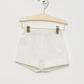A pair of Bonpoint Baby Girls' and Girls' Ecandy Shorts in White Poplin hang on a wooden hanger against a white background.