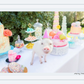 A piglet wearing a pink party hat stands on a table decorated with colorful cakes and sweets at a whimsical outdoor picnic, reminiscent of the Gray Malin Picnic Party II with its 29.5" x 41.5" white frame.