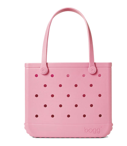 A pink, rectangular Baby Bogg® Bag tote from Bogg Bags features round holes on all sides and two handles. The word “bogg” is embossed on the lower right corner. This washable bag is ideal for everyday adventures and can be easily customized to reflect your unique style.