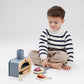 A child wearing a striped sweater engages in pretend play, handling toy pizza slices and pizza toppings from the "Tenderleaf Make Me a Pizza!" by Tender Leaf Toys with a wooden pizza oven toy, seated on the floor against a plain background.