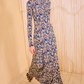 A person wearing the Ulla Johnson Naia Midi Dress with long sleeves and an ikat print in blue, beige, and black stands in a room with textured beige walls and floor.