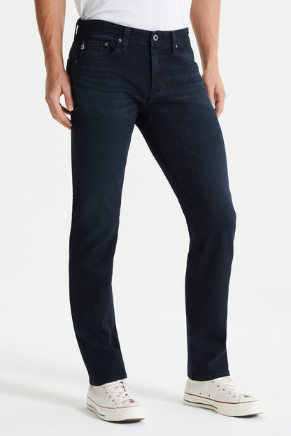 A person stands against a plain background, wearing AG Jeans' Everett Denim, Bundled in a dark wash, paired with a white t-shirt and pristine white sneakers.