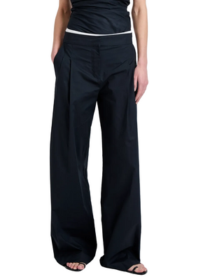 Someone wearing the Proenza Schouler White Label Coralie Pant—loose-fitting, high-waisted, wide-leg with front pleats and pockets—paired with a black top and open-toe black sandals.