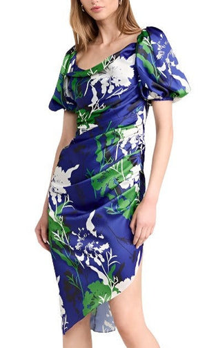 A woman wearing the Prabal Gurung Cowl Neck Side Ruched Dress, which is blue with green and white floral patterns, featuring short puff sleeves, a cowl neckline, and an asymmetrical hemline.