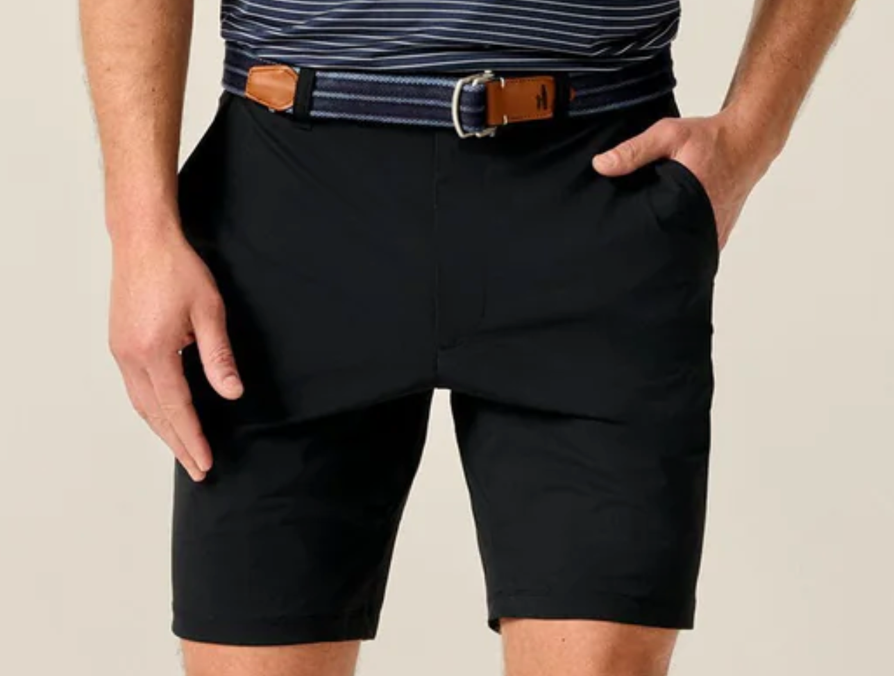 A person wearing a striped belt, a navy stretch-knit striped shirt, and Johnnie-O Fusionn Shorts in black with hands in pockets, shown from waist to above the knees.