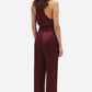 A person stands facing away, wearing a burgundy satin jumpsuit with wide-leg Novalis Fox Pants by Blaze Milano and black heeled shoes.