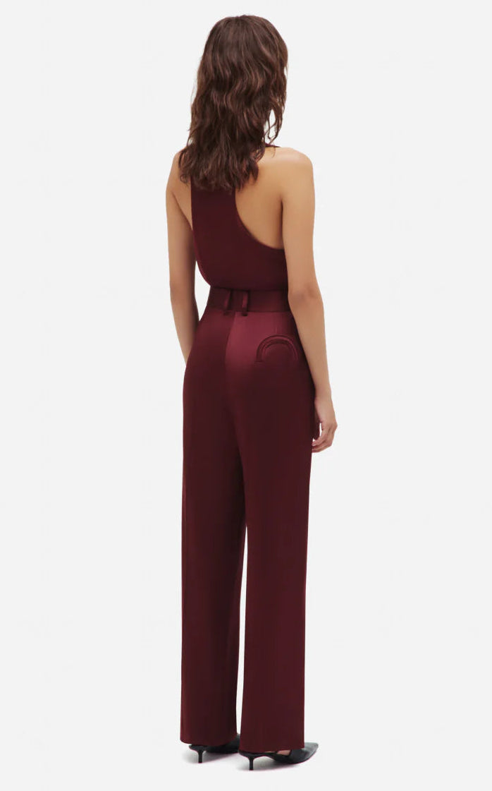 A person stands facing away, wearing a burgundy satin jumpsuit with wide-leg Novalis Fox Pants by Blaze Milano and black heeled shoes.