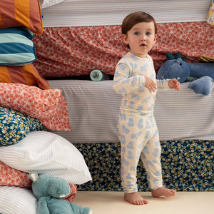 A toddler wearing the Magnetic Me Baby Boys' and Boys' Pudgemuffin Magnetic PJ Set stands on the floor beside a bed piled with colorful sheets, pillows, and stuffed animals.