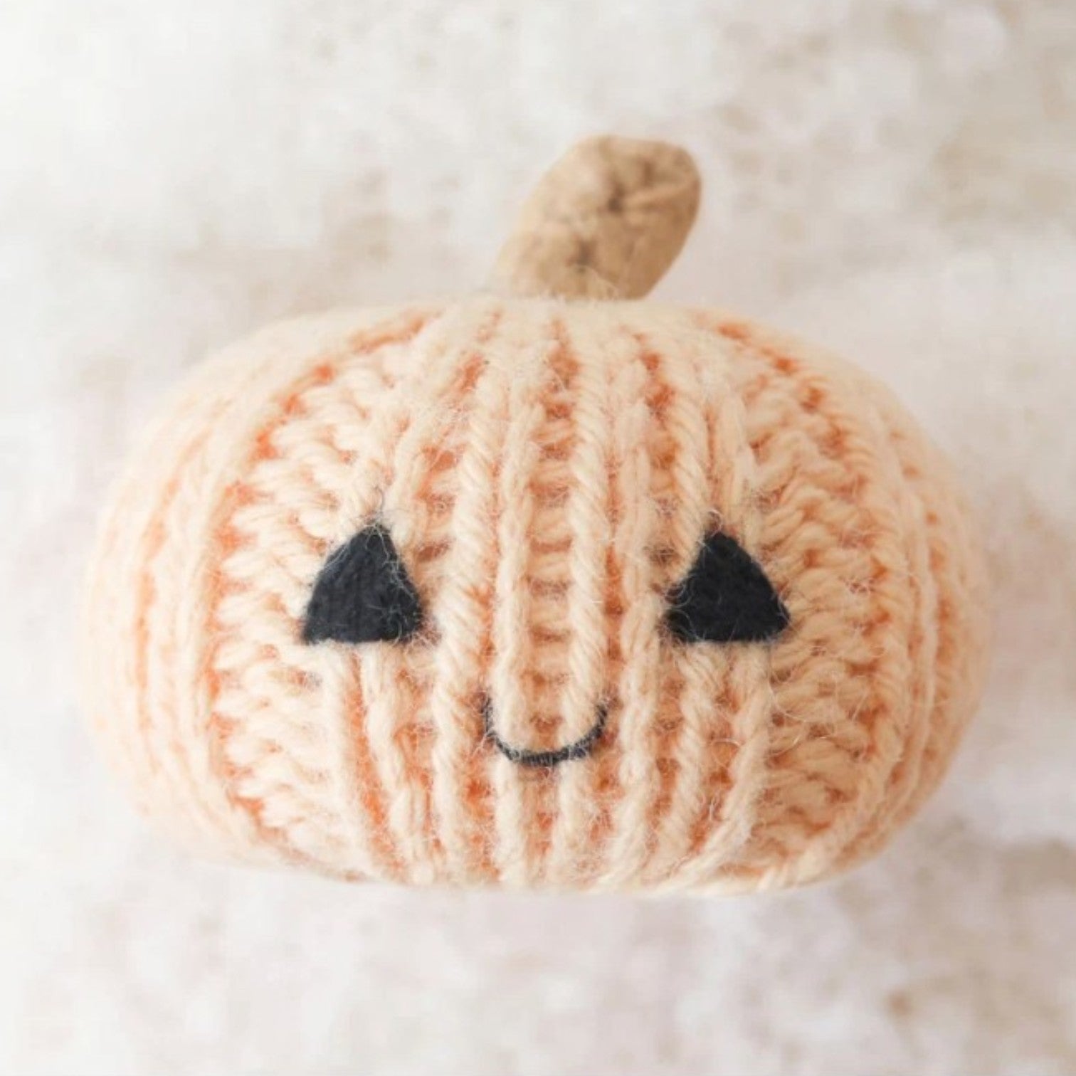 A small, knitted Baby Ghost - Pumpkin by Cuddle + Kind, with a beige stem and a simple face featuring black triangle eyes and a smiling mouth, sits on a white surface. Lovingly handcrafted from Peruvian cotton, this charming piece brings warmth and character to any space.