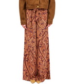 A person wearing Ulla Johnson’s Sawyer Silk Pant, featuring a red and orange paisley pattern in silk crepe de chine, paired with a brown jacket and standing against a plain background.