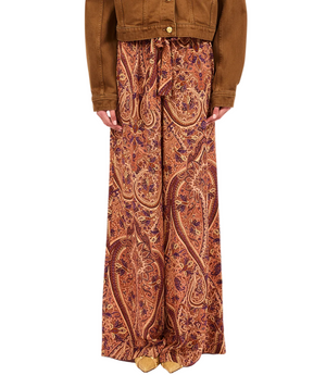 A person wearing Ulla Johnson’s Sawyer Silk Pant, featuring a red and orange paisley pattern in silk crepe de chine, paired with a brown jacket and standing against a plain background.