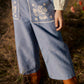 A child stands on dry grass, wearing the Louise Misha Girls’ Flofi Pants—light blue denim with white floral embroidery on the back pockets—paired with a cream-colored top and brown boots.