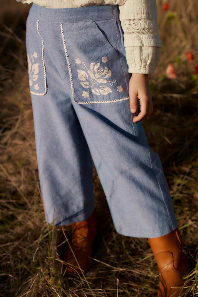A child stands on dry grass, wearing the Louise Misha Girls’ Flofi Pants—light blue denim with white floral embroidery on the back pockets—paired with a cream-colored top and brown boots.