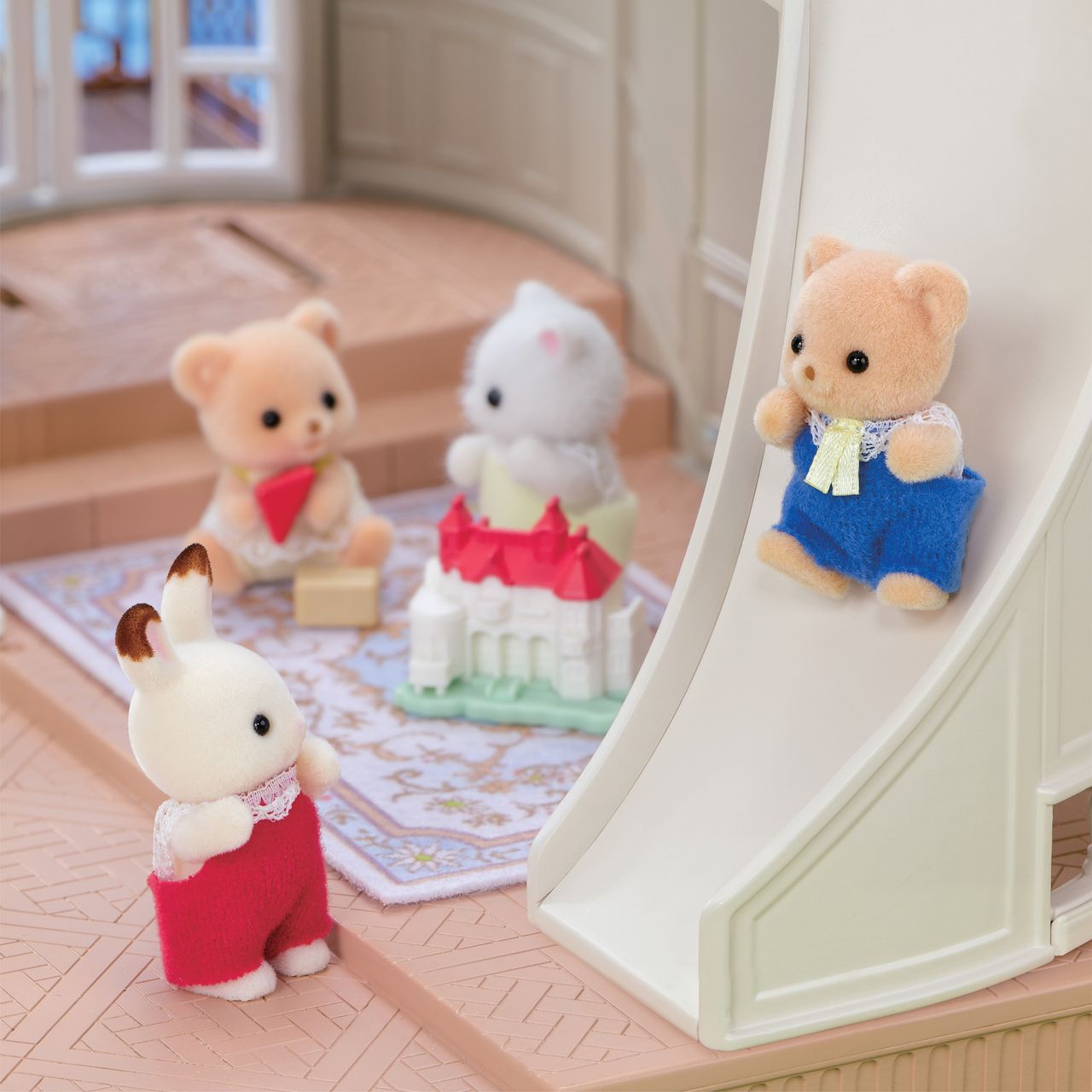 Four Calico Critters animal figurines play indoors; one bear in blue overalls slides down the spiral staircase slide of the Red Roof Riverside Manor while others sit or stand nearby next to the castle-style house on a patterned mat.