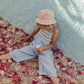 A child wearing Louise Misha's Morli Overalls and a floral hat sits on a petal-strewn cobblestone path, holding ice cream against a light blue wall. The scene is enhanced by textured fabric, offering a charming contrast with cotton voile accents.