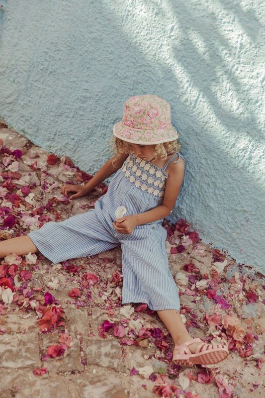 A child wearing Louise Misha's Morli Overalls and a floral hat sits on a petal-strewn cobblestone path, holding ice cream against a light blue wall. The scene is enhanced by textured fabric, offering a charming contrast with cotton voile accents.