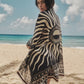 A person stands on a sandy beach facing the camera, wearing the Johanna Ortiz Golden Horizon Pareo. The black and white sun design contrasts with the ocean and partly cloudy sky, emphasizing their unique style.