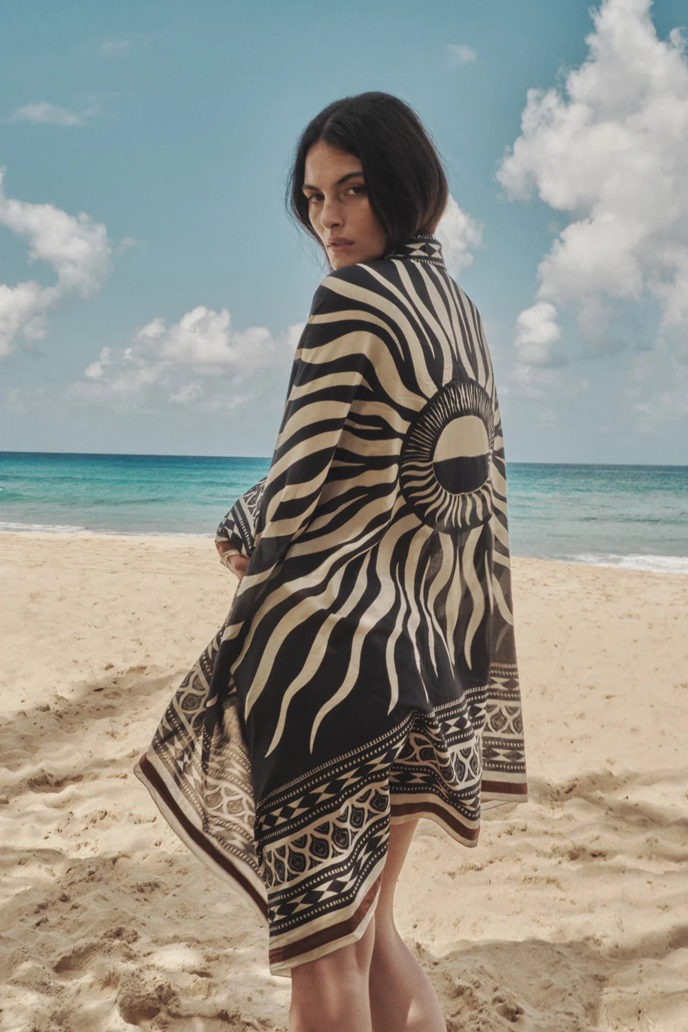 A person stands on a sandy beach facing the camera, wearing the Johanna Ortiz Golden Horizon Pareo. The black and white sun design contrasts with the ocean and partly cloudy sky, emphasizing their unique style.