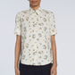 A person wears the Adam Lippes Charlie Top, a cream-colored, short-sleeve shirt with a collared design and a delicate floral and dinosaur pattern, standing against a plain background.