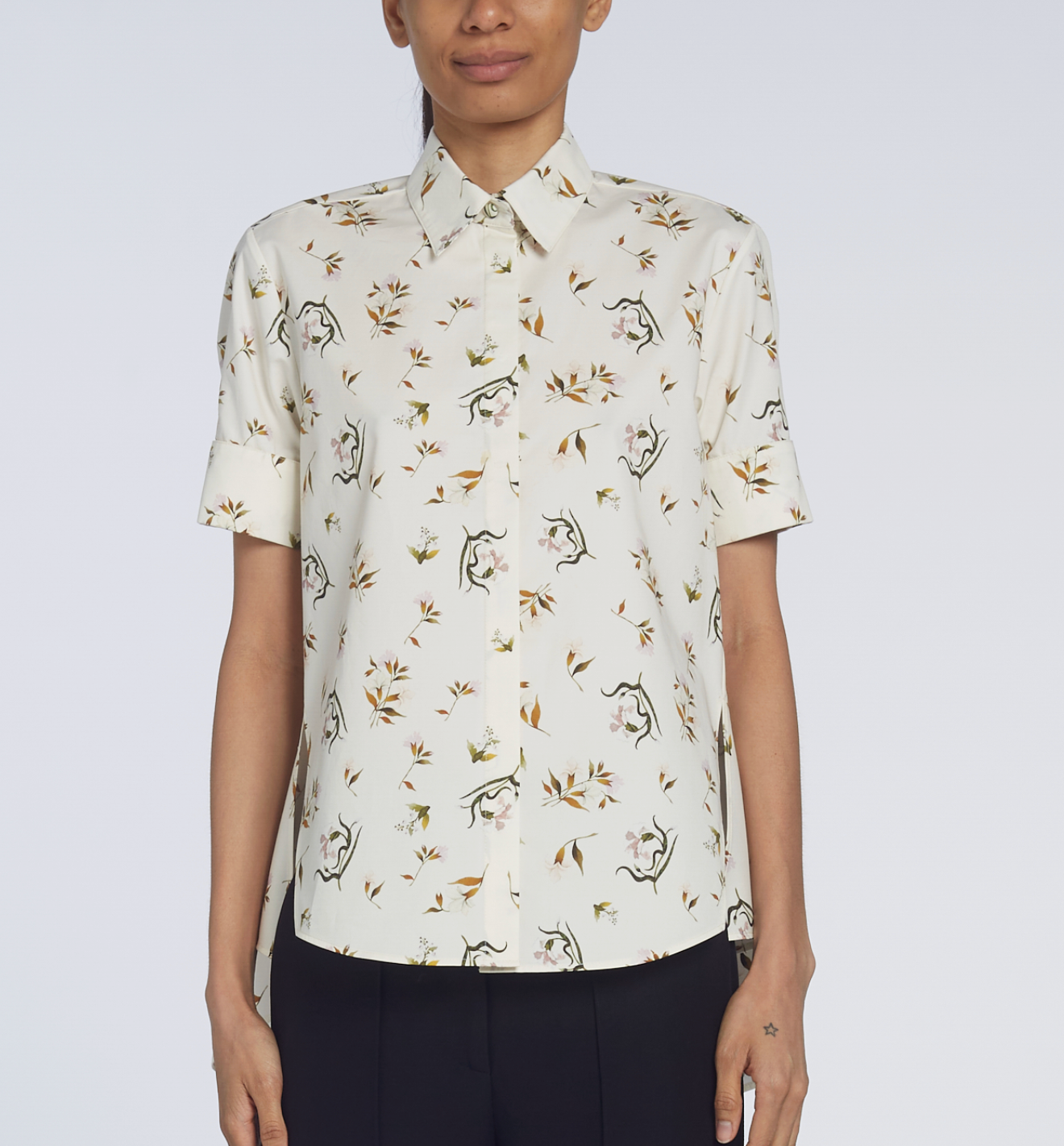 A person wears the Adam Lippes Charlie Top, a cream-colored, short-sleeve shirt with a collared design and a delicate floral and dinosaur pattern, standing against a plain background.