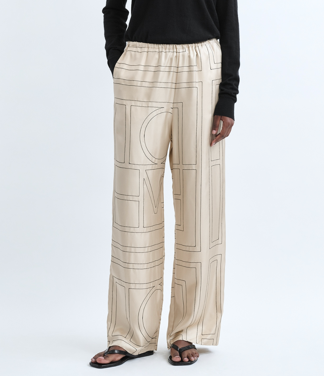 A person wears cream-colored Toteme Monogram Silk PJ Bottoms with black graphic lines, paired with a black long-sleeve top and black sandals, standing against a plain background.