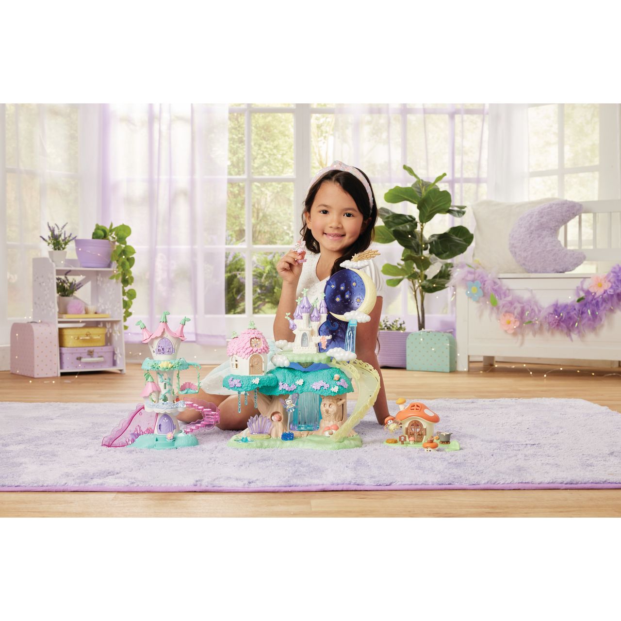 A young girl sits on a purple rug, smiling and playing with the Calico Critters Woodland Wonder Fairy Castle and enchanting fairy accessories in a brightly lit room with plants and pastel decor.
