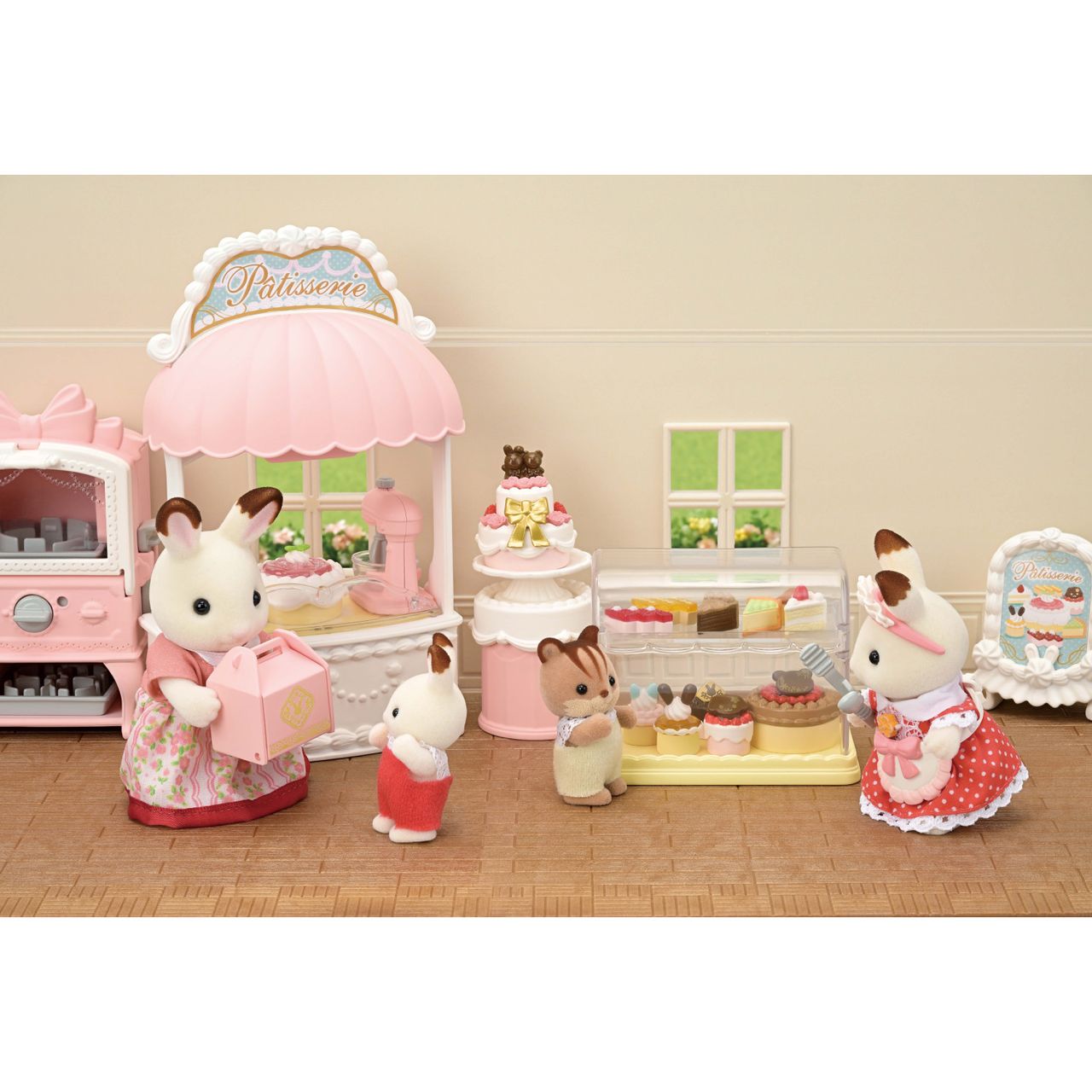 The Calico Critters Village Cake Shop Starter Set includes four animal figures in a pink cake shop, featuring cakes on display, a cake baking in the oven, and one figure holding a pink box by the pastry counter decorated with delightful cake accents.