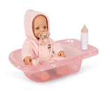 A Konges Slojd Isola Doll Bath Set featuring a baby doll in a pink hooded bathrobe and pacifier sitting in a translucent pink plastic bathtub next to a toy bottle.