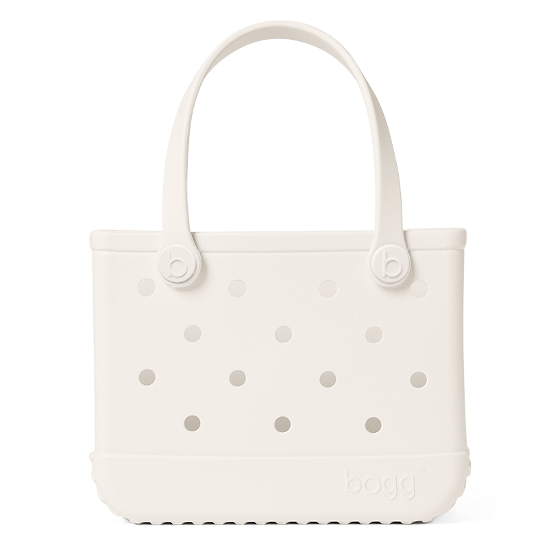 The Bitty Bogg® Bag by Bogg Bags is a white, rectangular tote featuring round side holes, sturdy handles, and the “bogg” logo embossed near the bottom corner. This compact tote is ideal for carrying daily essentials, and its washable design ensures easy cleaning and long-lasting durability.