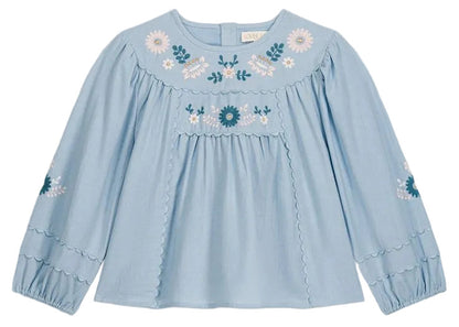 The Girls’ Risa Blouse by Louise Misha is crafted from light blue cotton and linen, adorned with floral embroidery on the chest and sleeves. It features scalloped trim, gathered cuffs, and subtly puffy sleeves for a delicate finish.