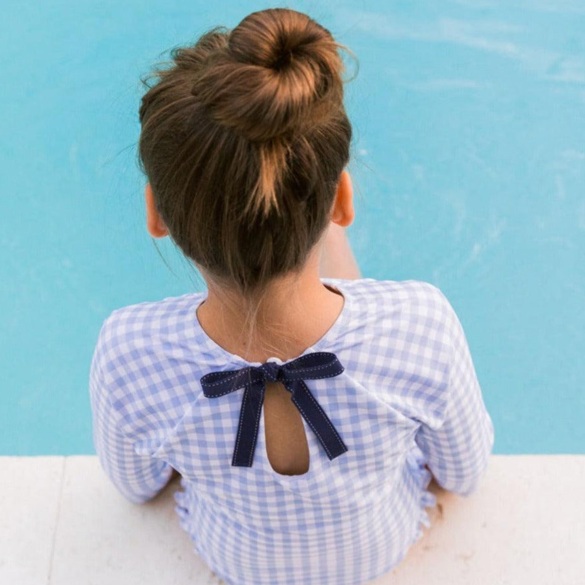 A girl with a bun hairstyle sits by a swimming pool, wearing the Minnow Girls' Rashguard One Piece by Minnow, which features a light blue and white checkered outfit with ruffle hip detail and a navy bow on the back.