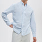 A person in a Rag & Bone Classic Oxford Shirt, light blue and white striped, long-sleeved, classic fit yarn-dyed cotton, pairs it with light-colored pants while standing against a plain background.