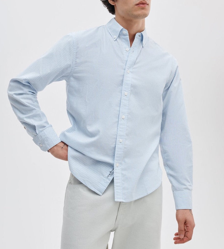 A person in a Rag & Bone Classic Oxford Shirt, light blue and white striped, long-sleeved, classic fit yarn-dyed cotton, pairs it with light-colored pants while standing against a plain background.