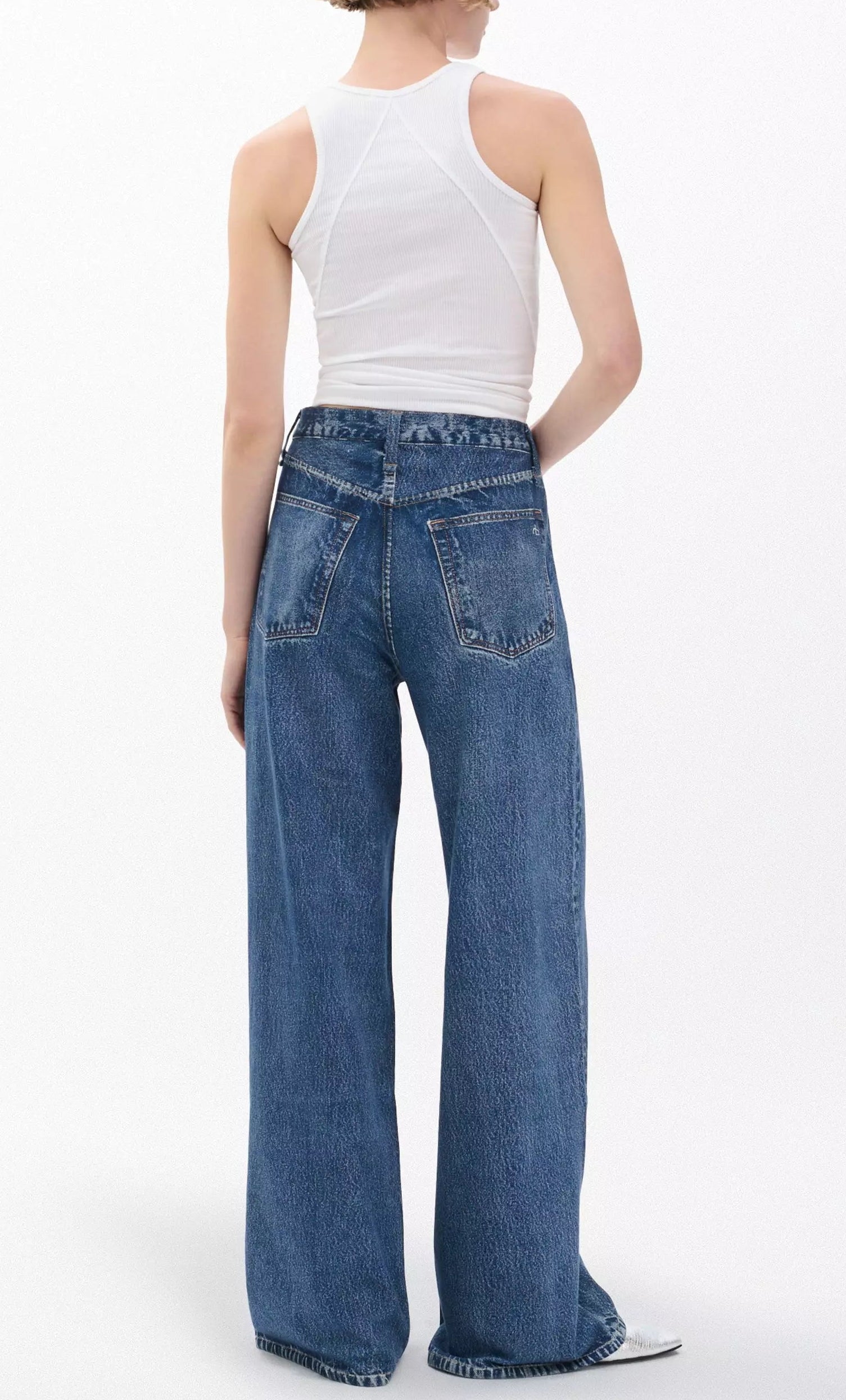 A person stands with their back to the camera against a plain light background, wearing a white sleeveless top and Rag & Bone Miramar Terry Sofie Full blue jeans.