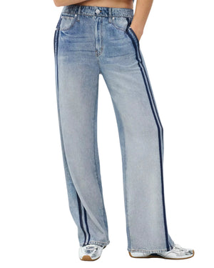 A person wears Rag & Bone's Miramar Terry Wide Track Pant—high-waisted, light-wash, wide-leg jeans with dark blue side stripes—paired with white sneakers and hands in pockets.