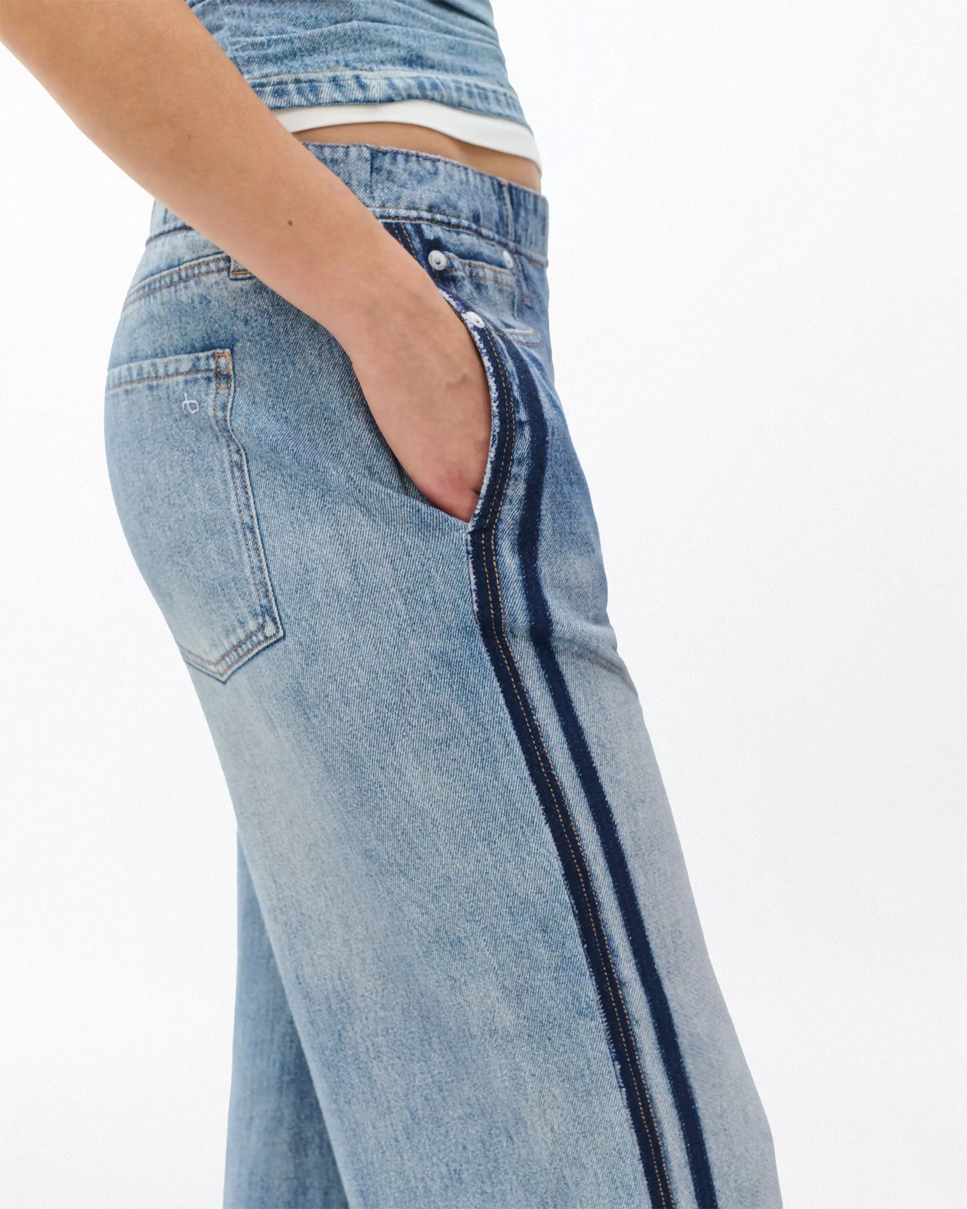 A person wearing Rag & Bone's Miramar Terry Wide Track Pant, featuring a wide-leg fit in light blue with dark blue side stripes, stands with one hand in their pocket against a plain white background.