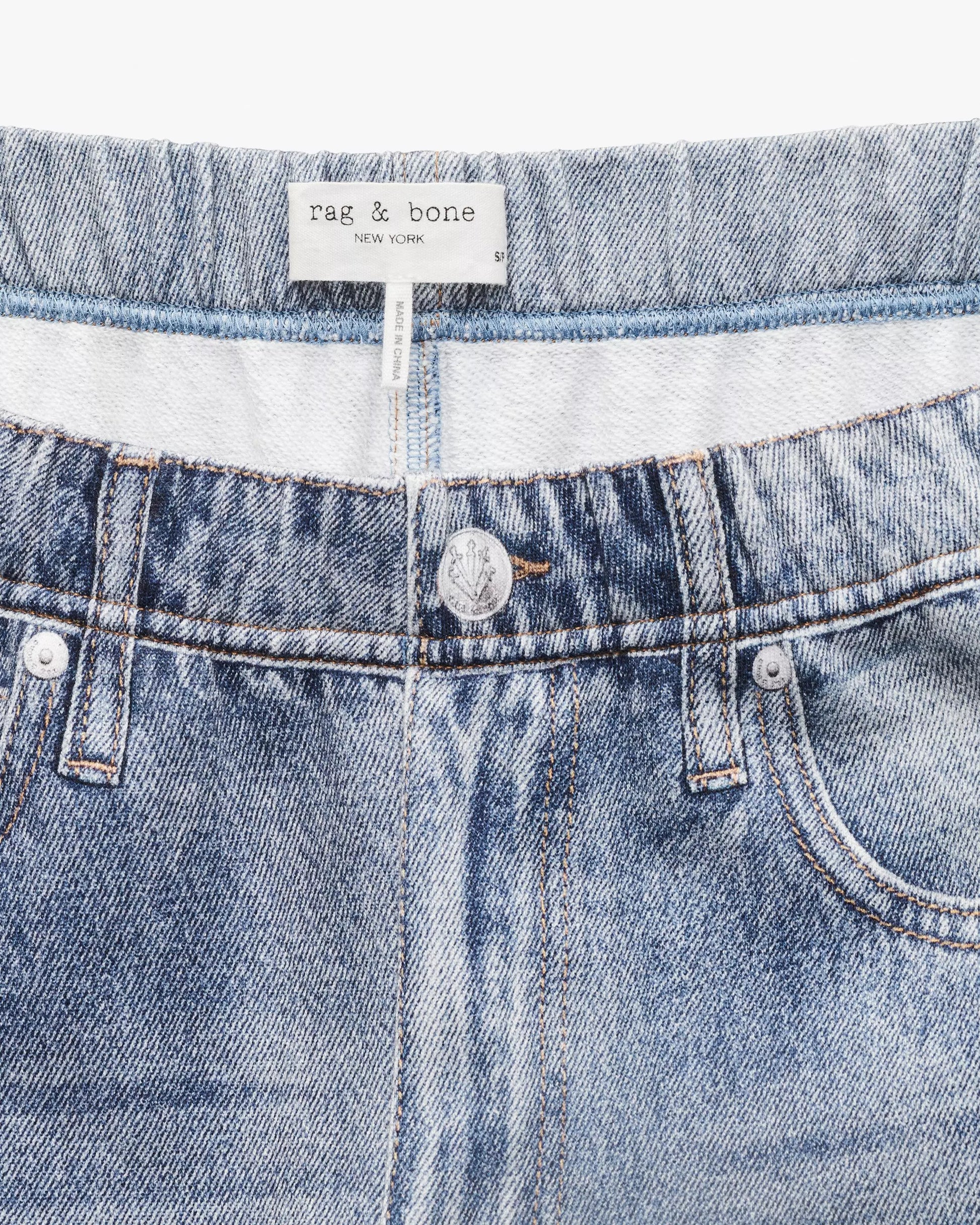 Close-up of the Miramar Terry Wide Track Pant by Rag & Bone, featuring blue denim-look fabric, silver button, pockets, a "rag & bone NEW YORK" label, and a hint of elastic waistband for comfort.