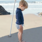 A toddler wearing a sun-safe, navy Minnow Unisex Rashguard and striped shorts stands on a beach under an umbrella, looking down at the sand. The ocean waves are visible in the background.