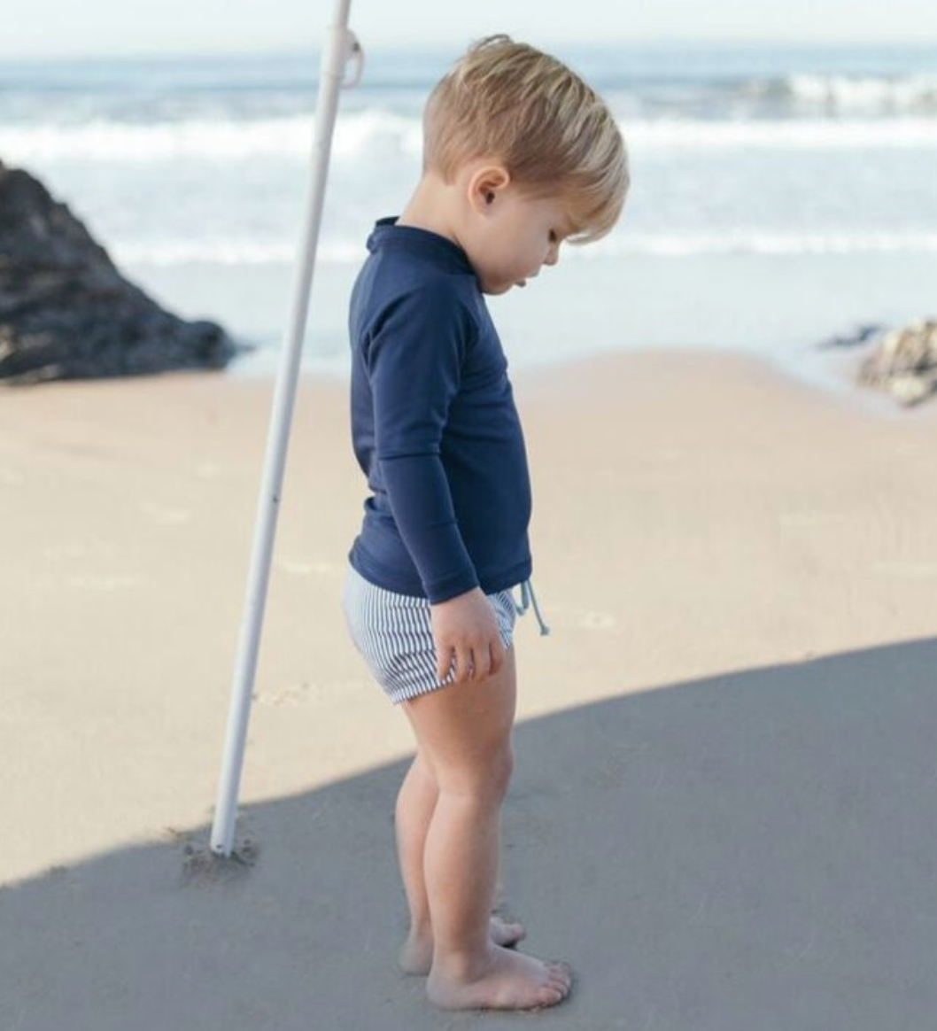 A toddler wearing a sun-safe, navy Minnow Unisex Rashguard and striped shorts stands on a beach under an umbrella, looking down at the sand. The ocean waves are visible in the background.